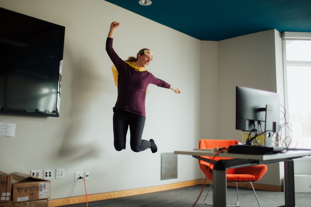 woman jumping in the air wearing a cape in front of  computer for virtual programming