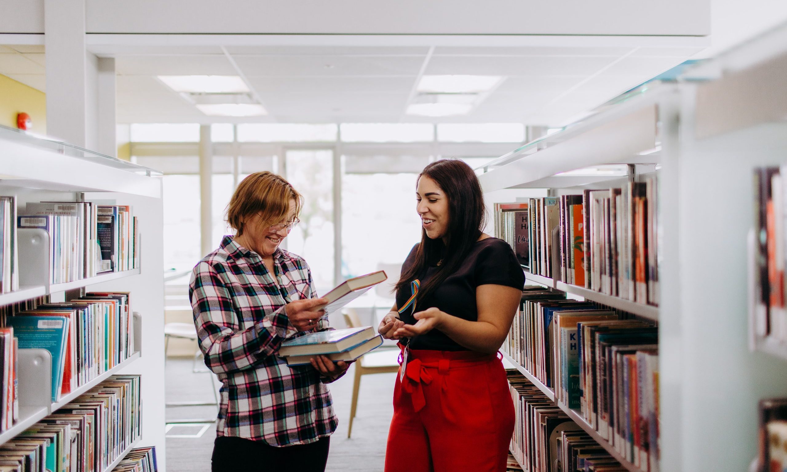 Library staff suggesting books to customer, two women standing in aisle of book shelves