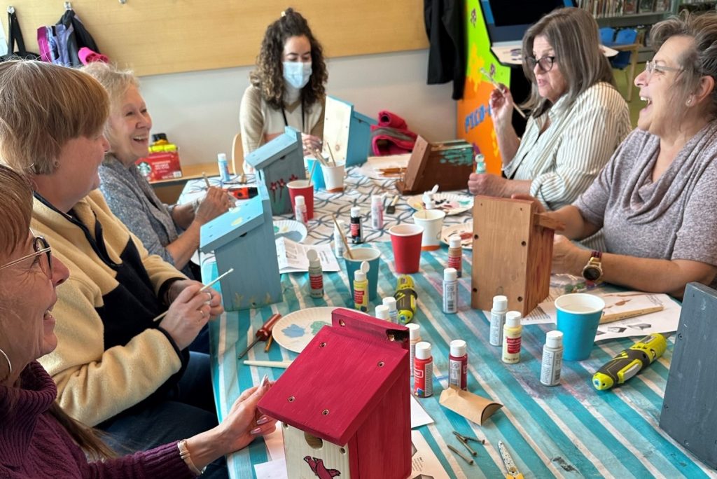 A group of senior citizens are smiling and laughing while painting birdhouses