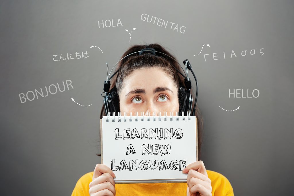 Portrait of a young woman wearing headphones, holding a notebook and looking up at a foreign greeting, covering half of her face. Gray background. The concept of learning foreign languages.